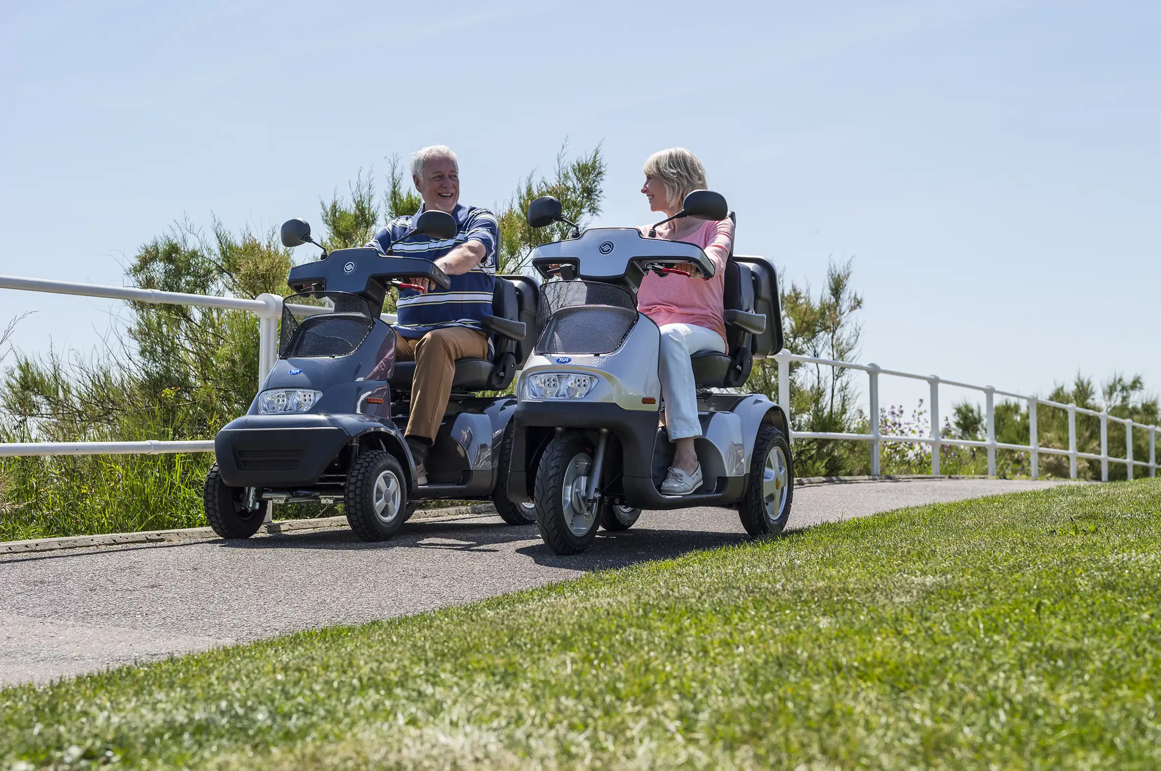 Two older adults riding mobility scooters side by side along a sunny seaside path